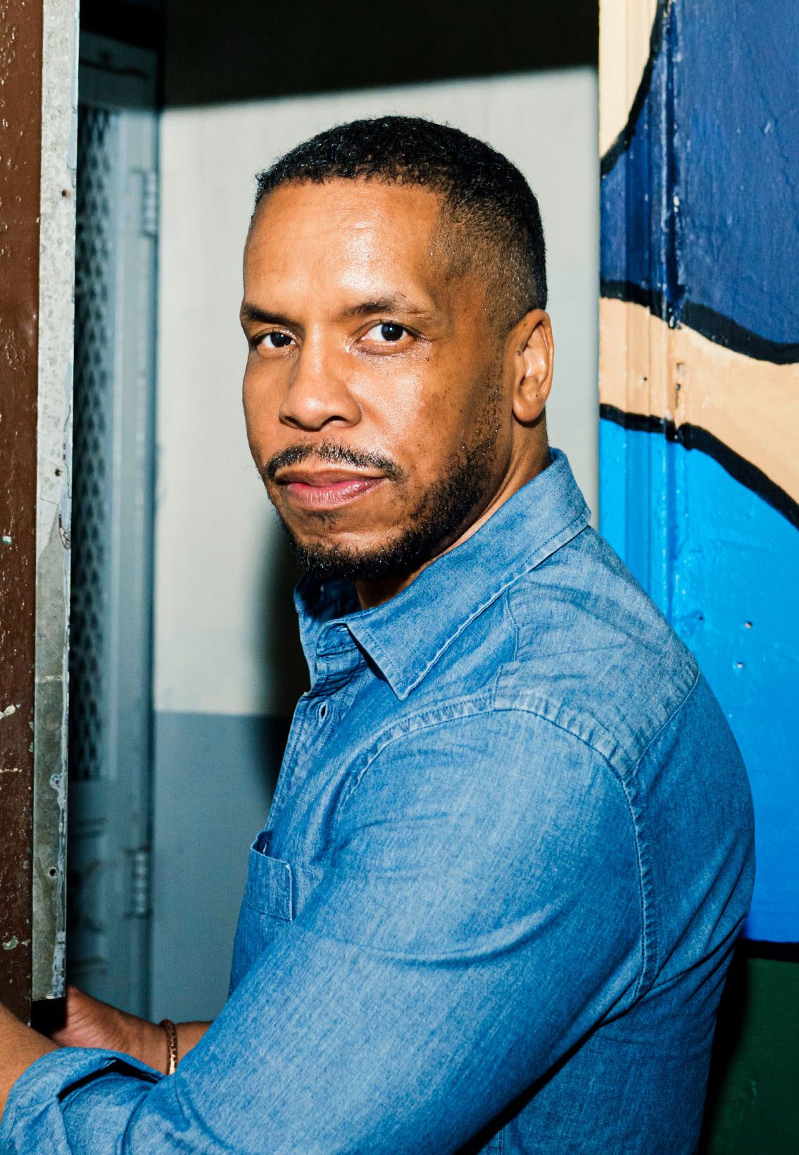 Gregory Purnell wearing a blue denim dress shirt, at the front door of his studio. Door is painted with art in multiple shades of blue, black, and beige.
