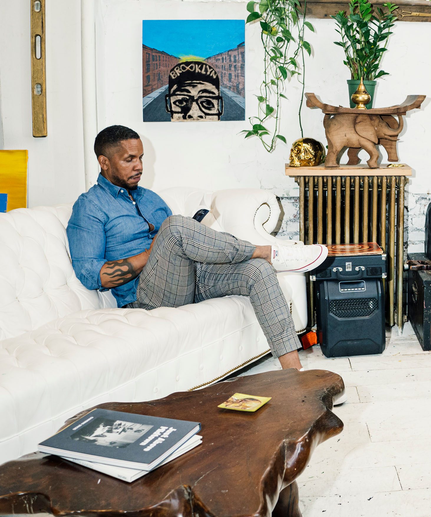 Gregory Purnell seated on a white couch in his studio. The space is decorated with plants, artwork, and a wooden table with a natural edge. A clerestory window runs along the top of the wall in the background, bringing in natural light.