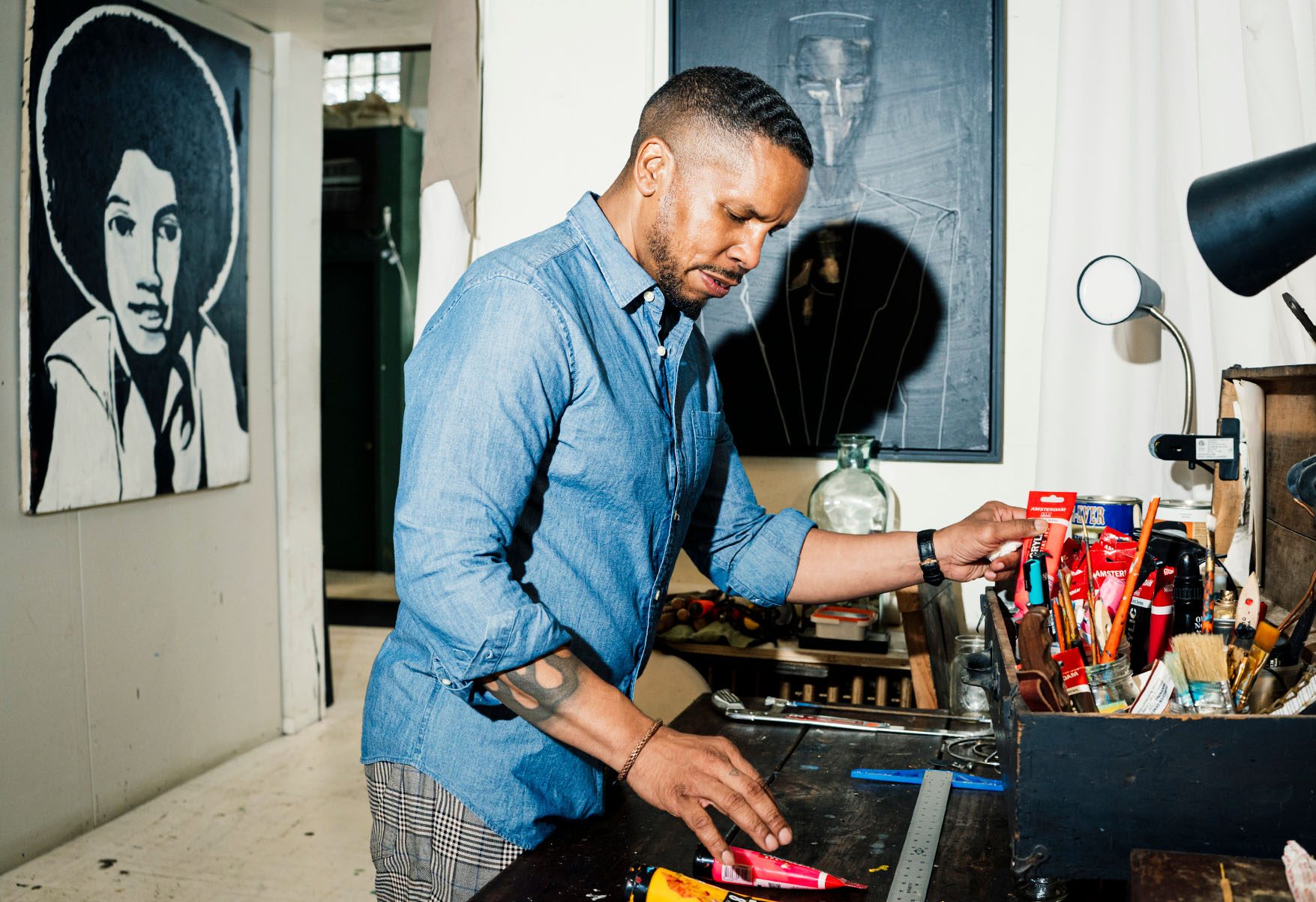 Gregory Purnell stands in his studio. In front of him is a desk with paint brushes and tubes of paint. Artwork decorates the walls of the space.