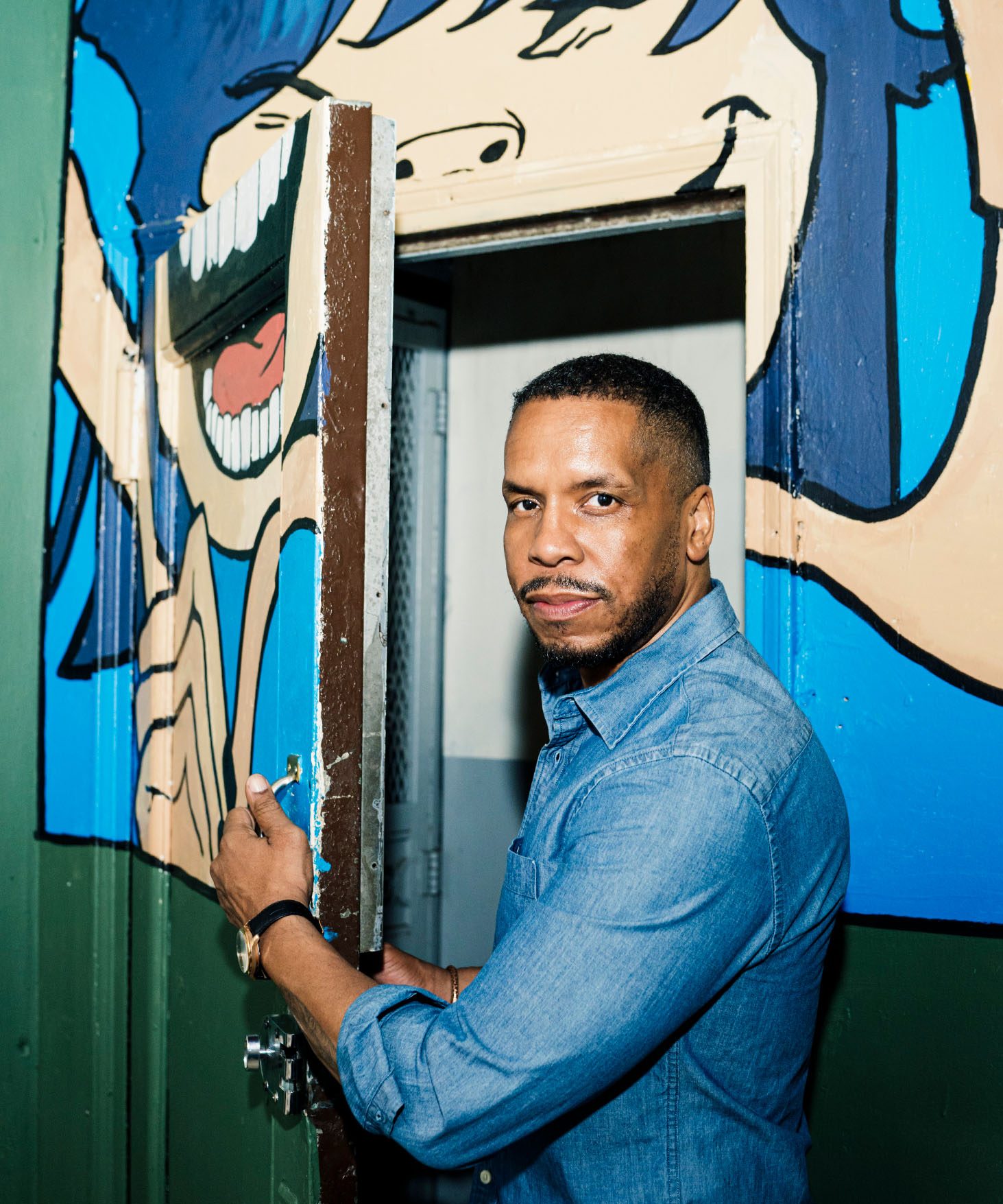 Gregory Purnell stands in front of the door to his studio where a large mural is painted of a person with blue hair, smiling.