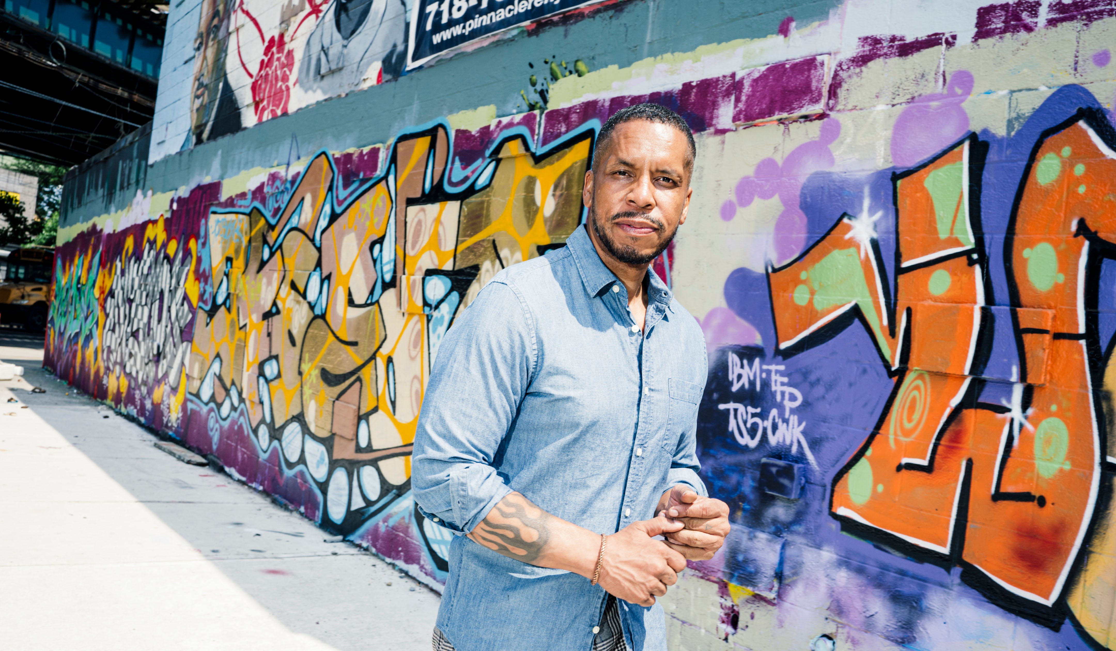Gregory Purnell stands in front of a long colorful graffiti wall near his studio in Brooklyn. In the background is an elevated subway track.