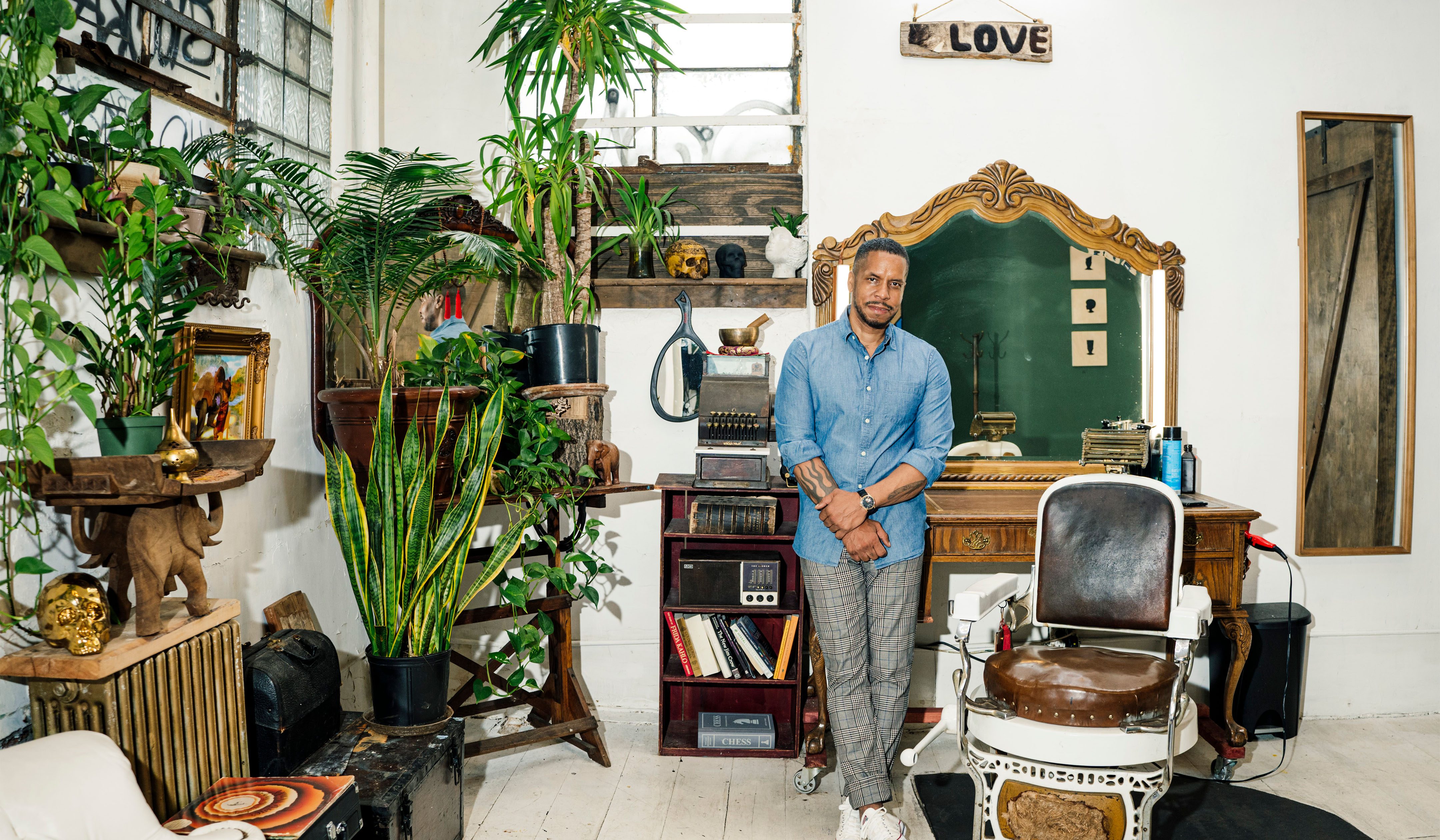 Gregory Purnell standing in his studio filled with various plants, sculptures, a barber's chair, and large mirror in the background.