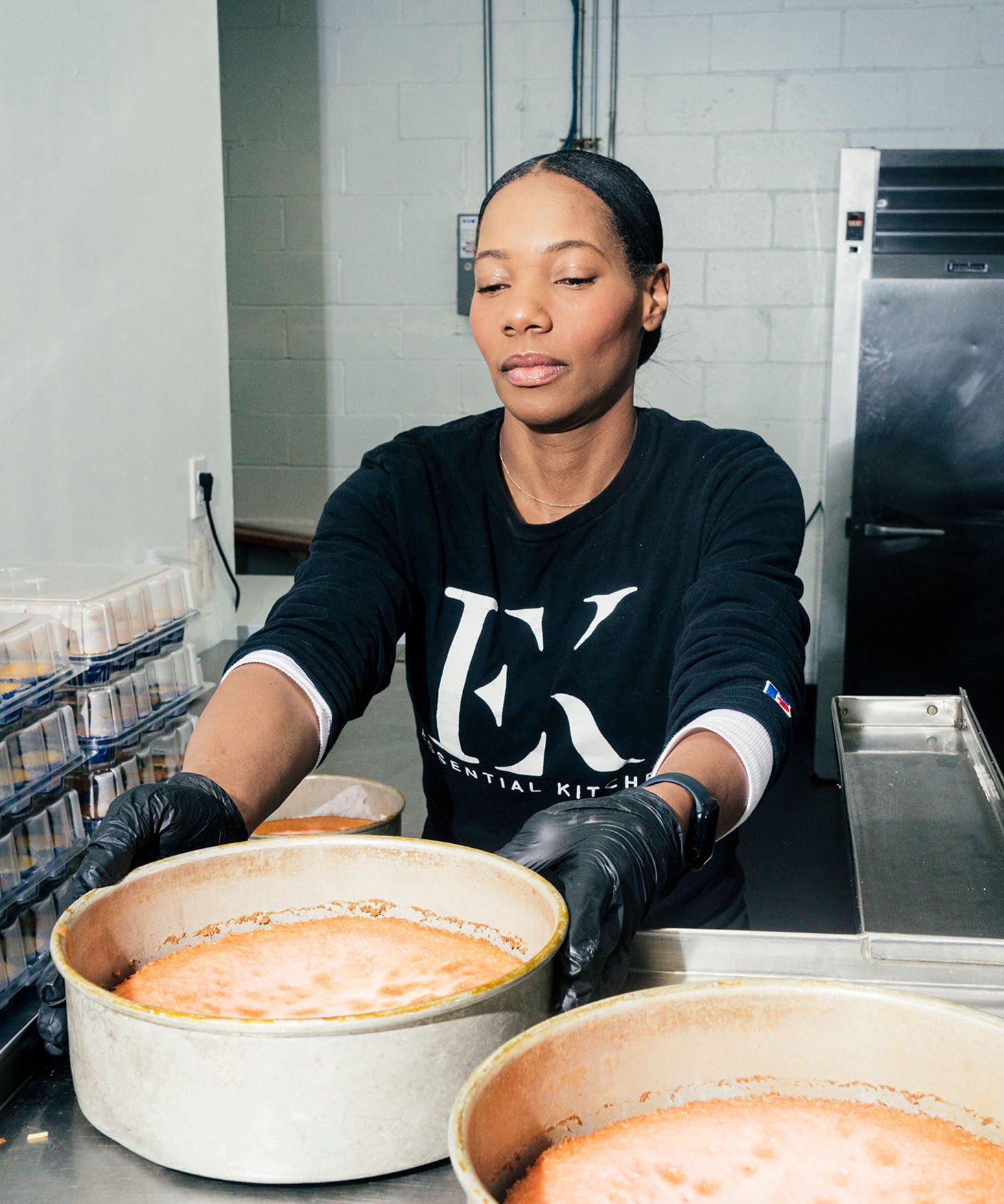 Dianna Rose, working in a kitchen, holding a large deep cake pan with cake visible inside. Dianna is wearing black gloves and a black long-sleeve shirt with large Essential Kitchen logo printed on it.