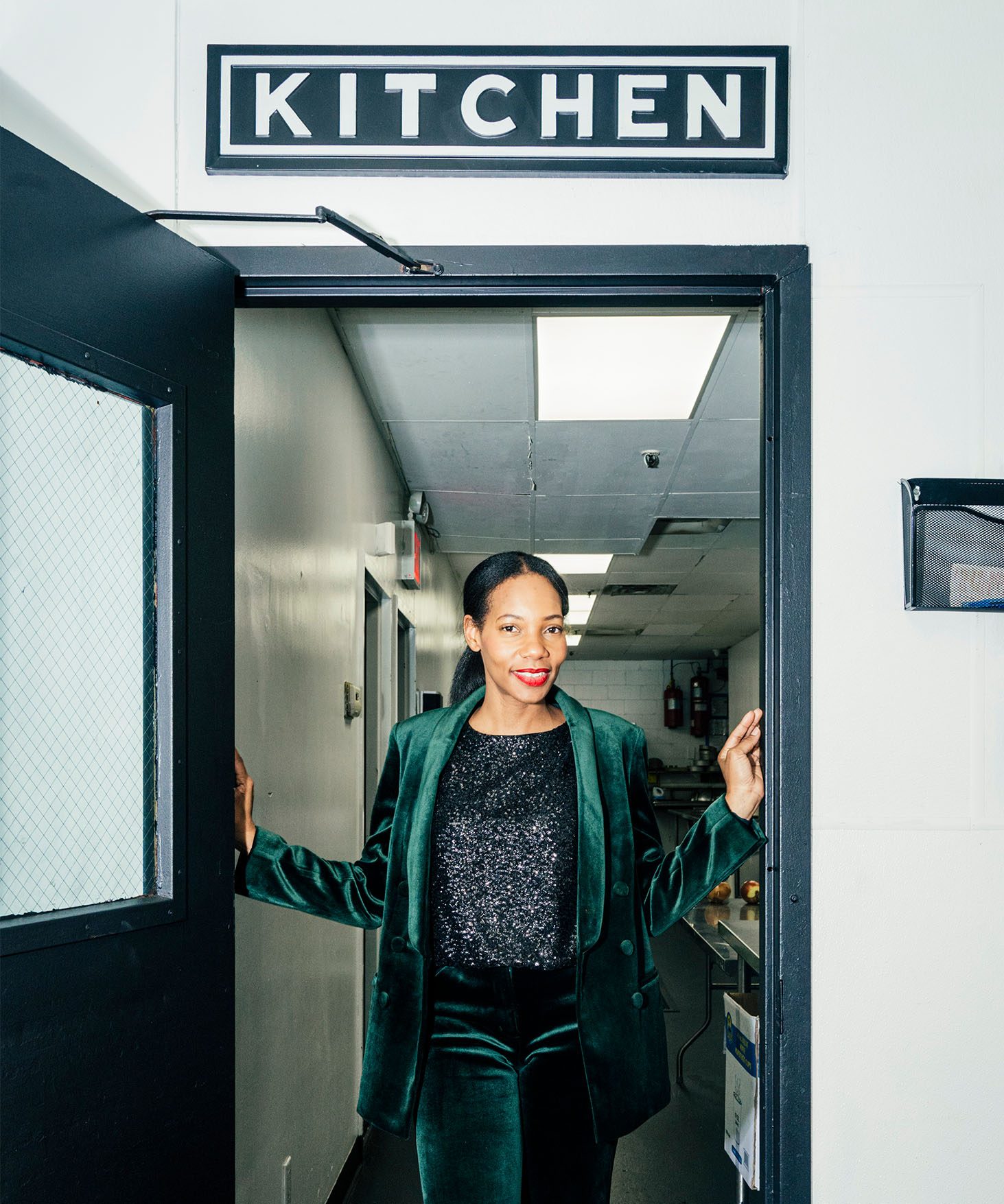 Dianna Rose standing in a doorway in green velvet suit. Directly above her is a sign that reads, “kitchen.”