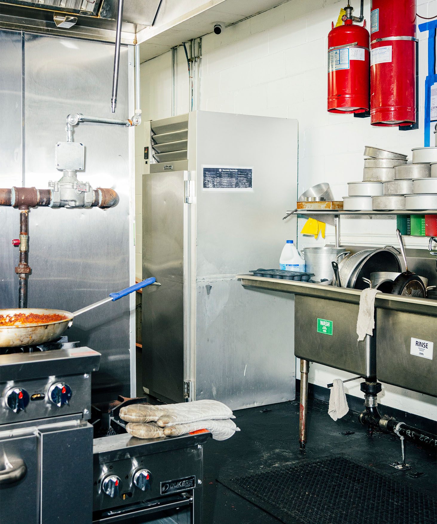 View of a large commercial kitchen. Professional stove is visible in front, with a stainless steel commercial dish sink full of dishes in the back.