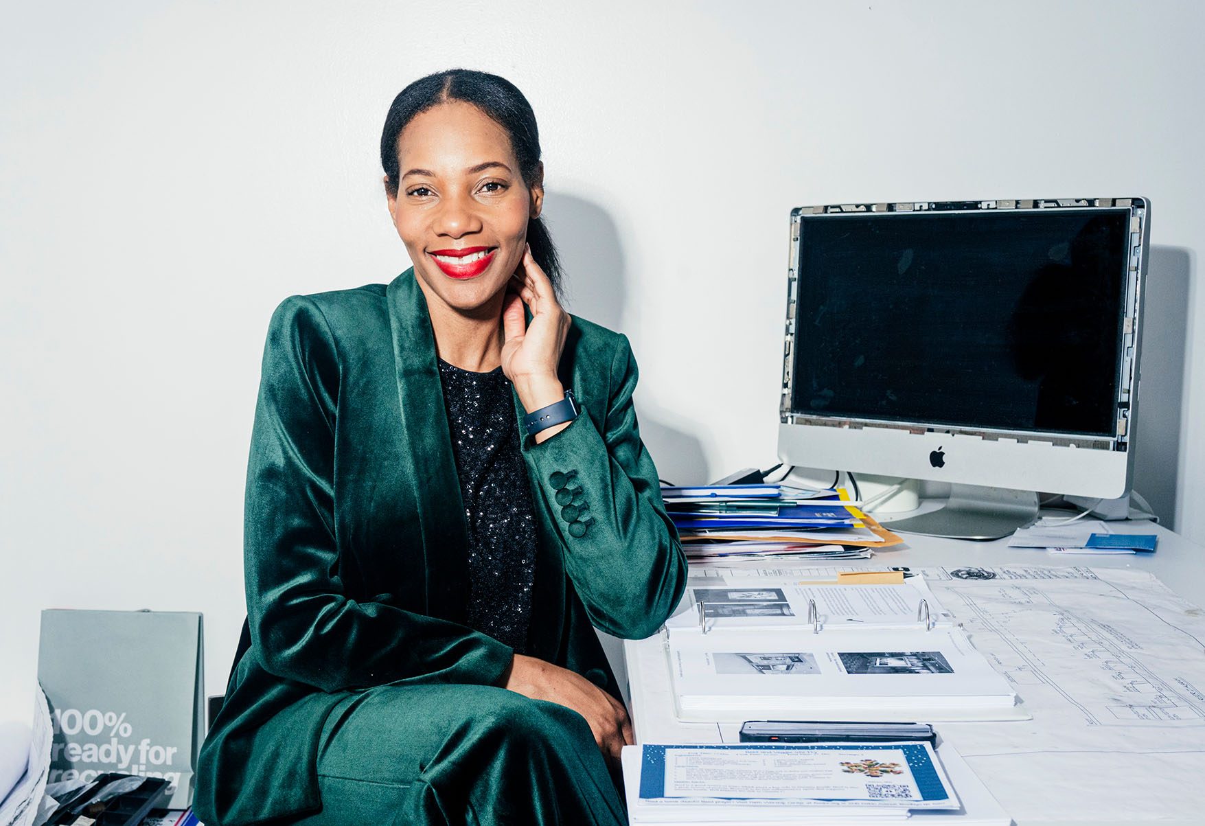 Dianna Rose smiling and seated at a work desk in an office with white walls. She is wearing a green velvet suit and an old, partially disassembled iMac computer is visible on the right.