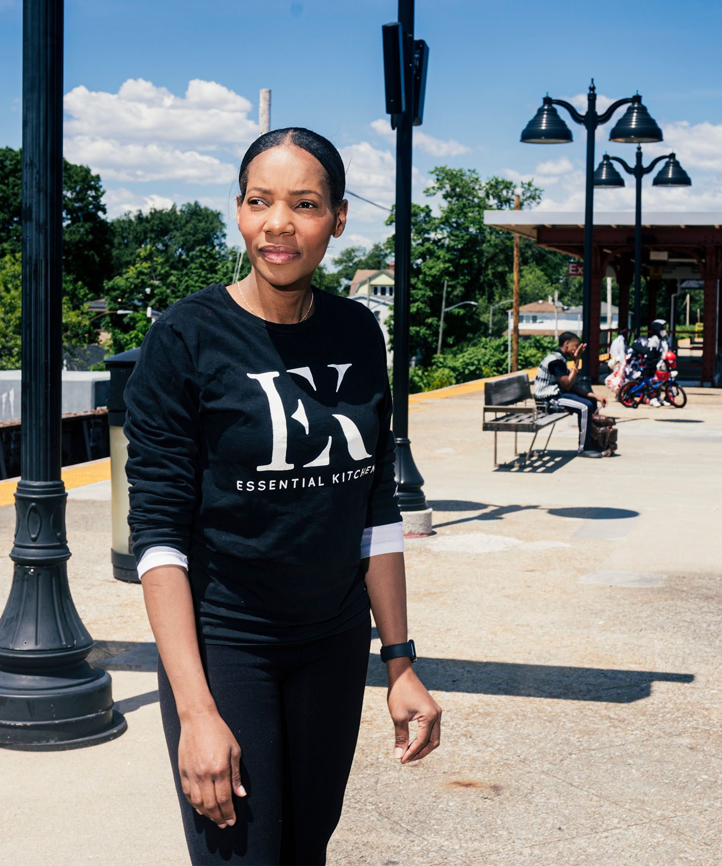 Dianna Rose, standing on an outdoor LIRR train platform in a bright sunlight. Covered waiting area is visible in the back. There are a few people seated on benches.