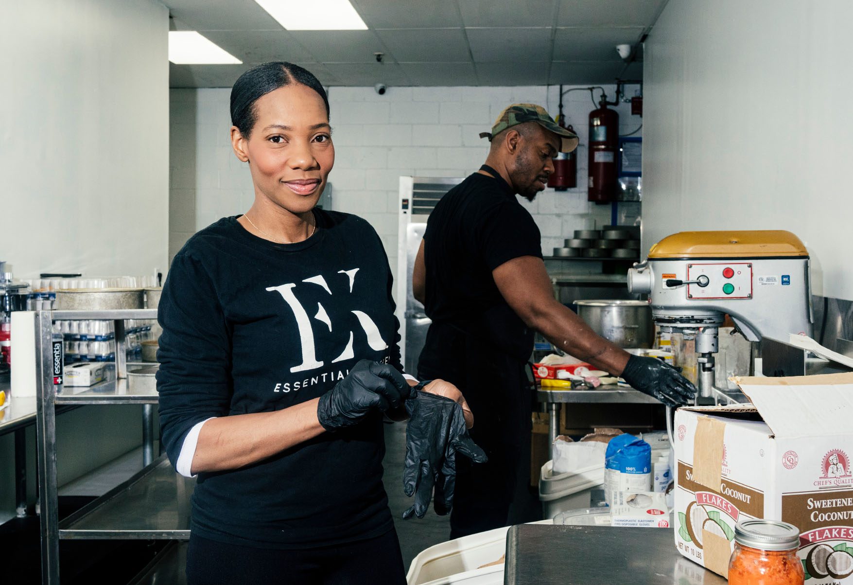 Dianna Rose putting on black work gloves facing the camera in a kitchen. In the background is another person in identical gloves, using a commercial food mixer.