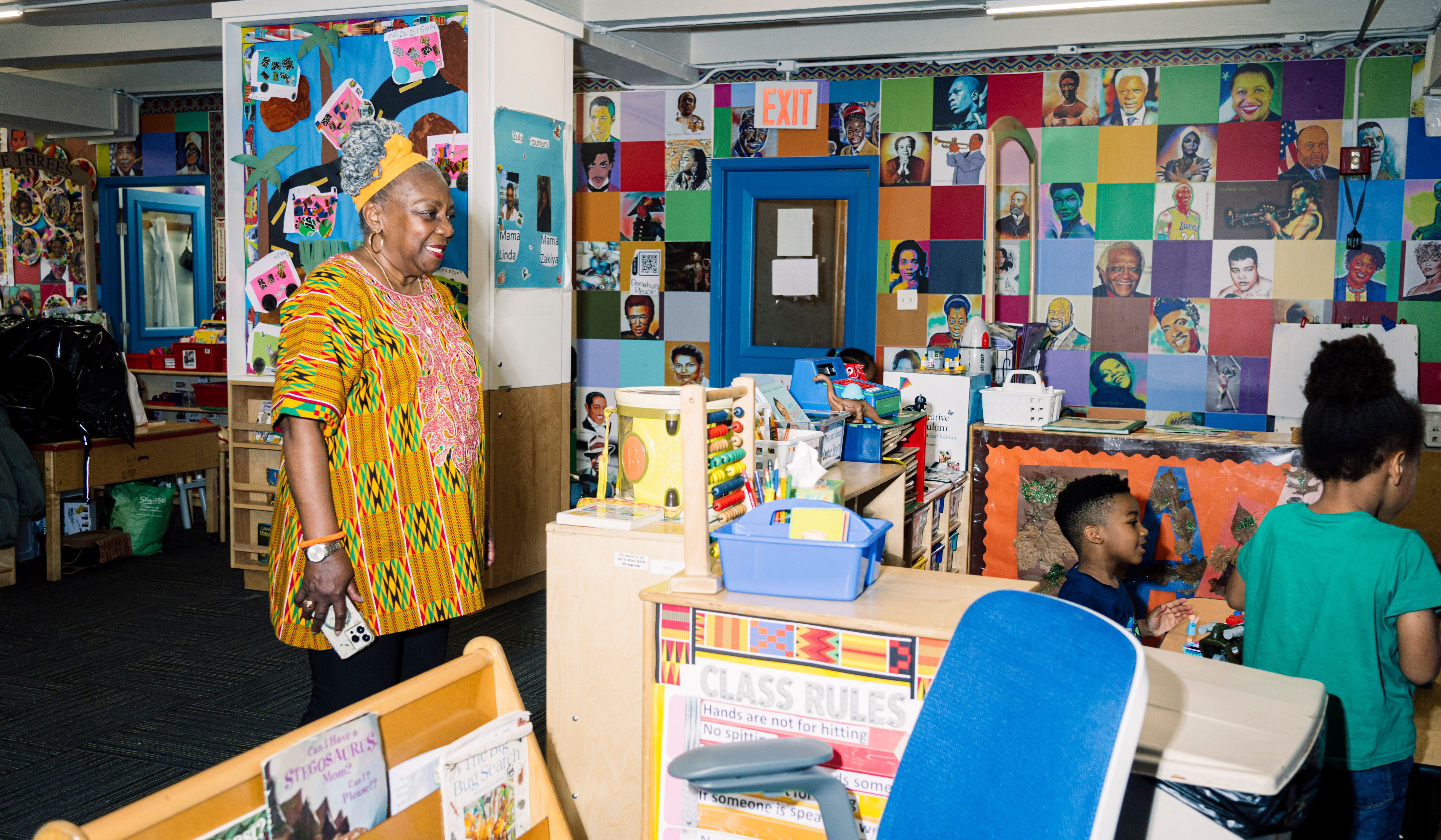 Fela Barclift stands and smiles in a colorful, decorated classroom watching kids having activities. In the background is a wall of artwork of popular figures. Tables with books and toys are in the center of the room.