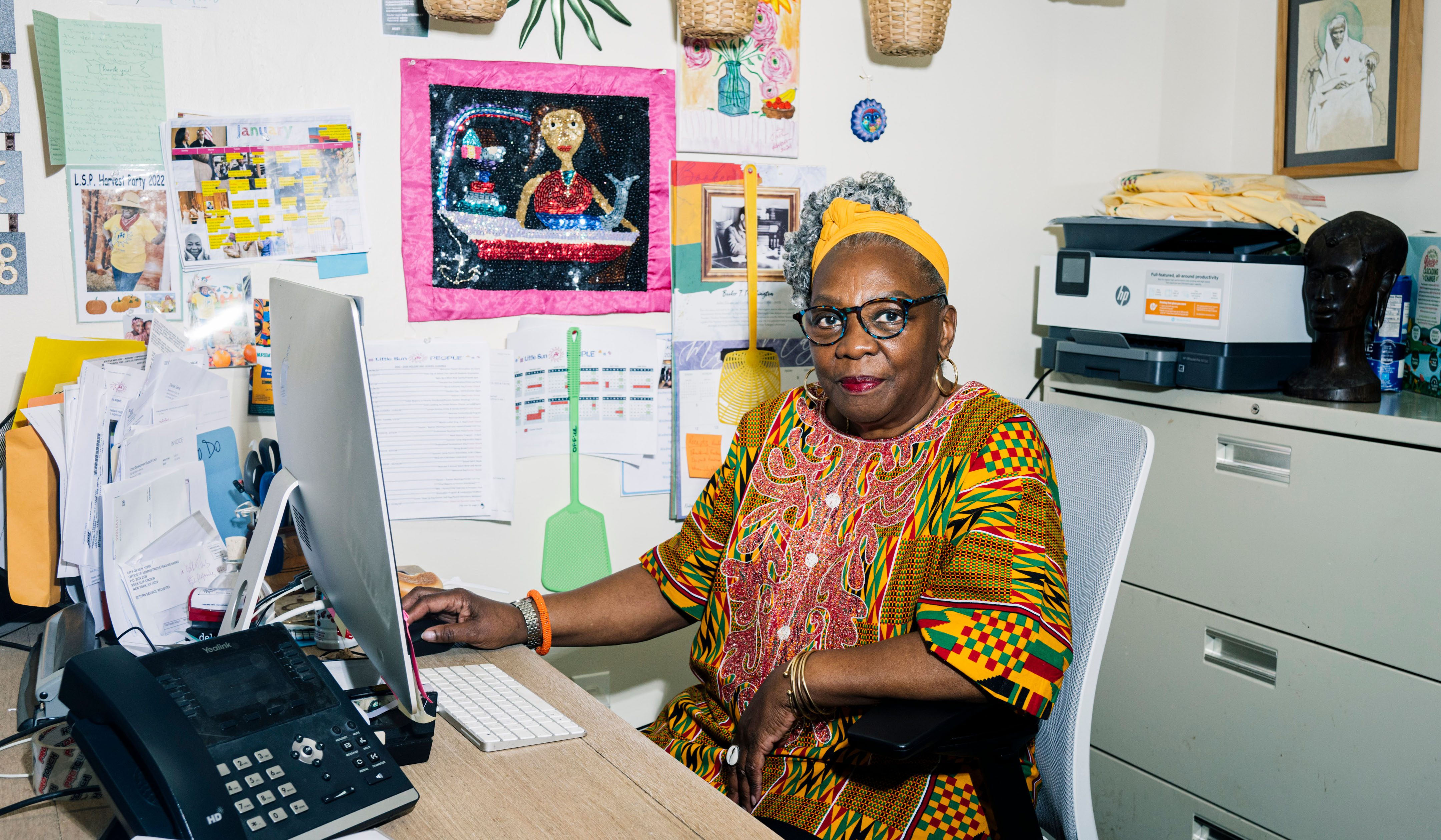 Fela Barclift, seated at an office in a school. She is in front of an iMac computer with a printer behind her. The wall behind her is adorned with magazine clippings, photos, drawings, and two plastic fly swatters.