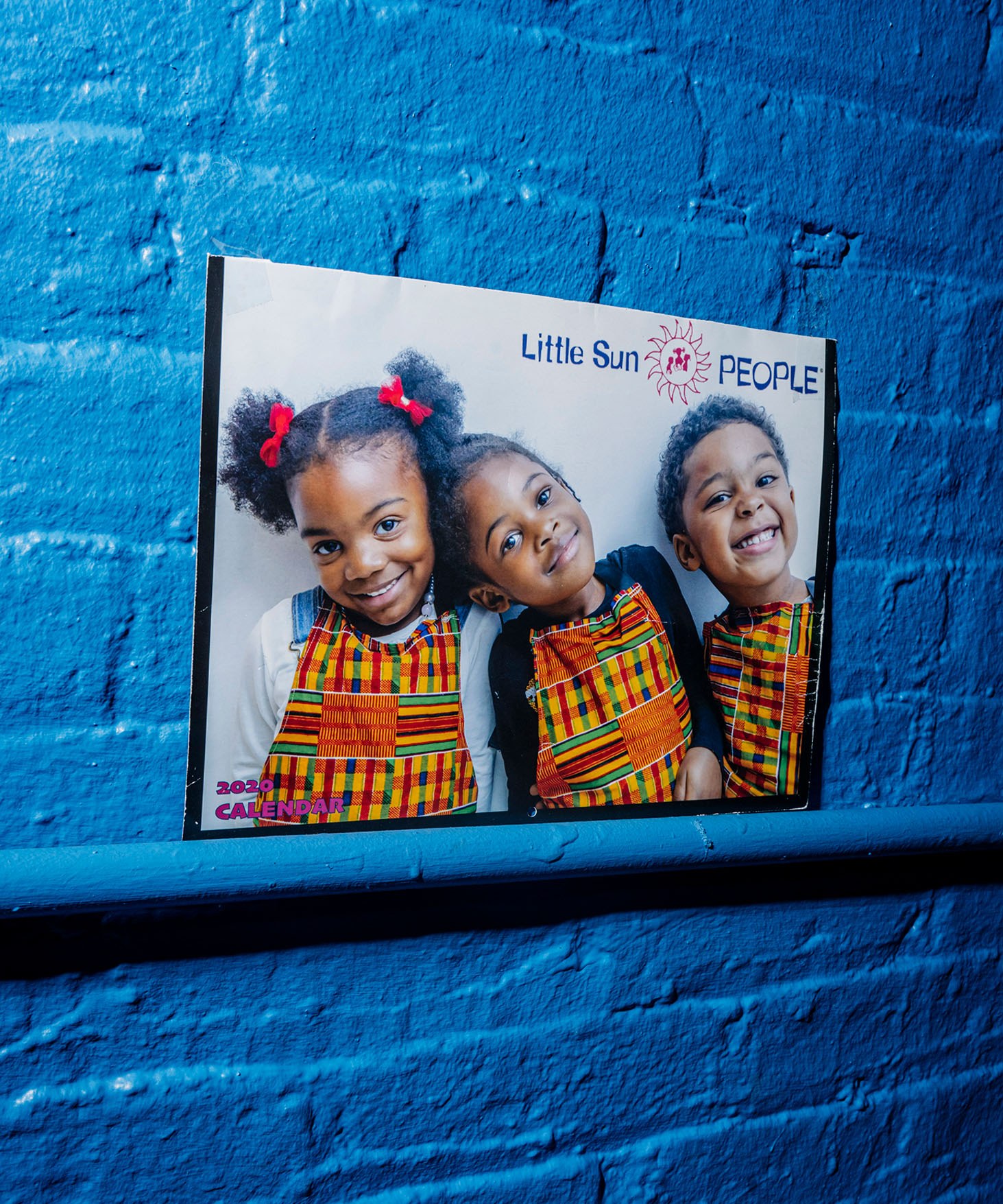 A framed photo on a bright blue wall with three children smiling and leaning on each other. At the top is the logo for Little Sun People.