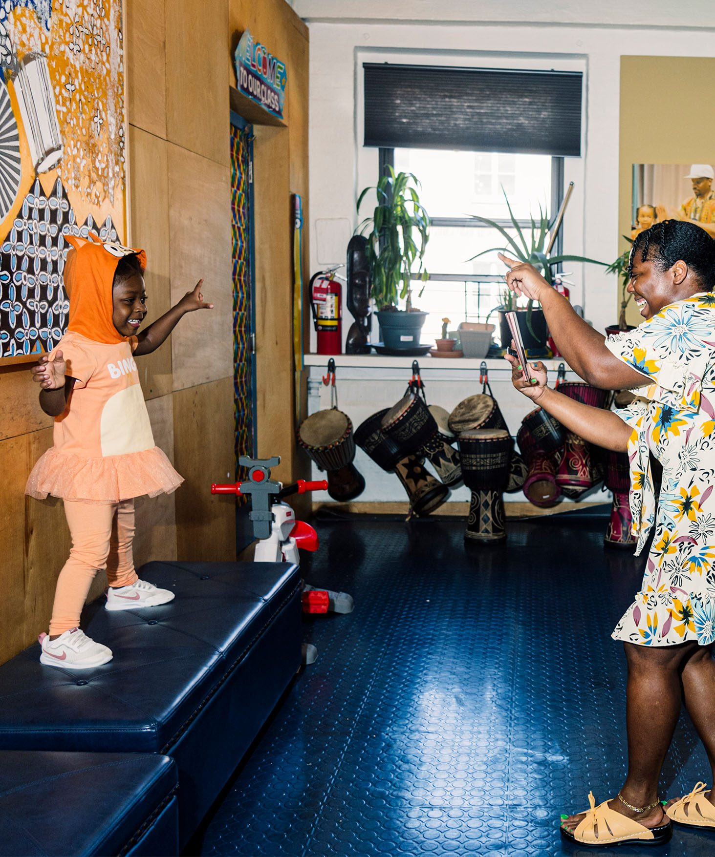View of a child in costume, dancing on a small stage in a classroom as an adult takes their photo. In the background are a row of African drums.