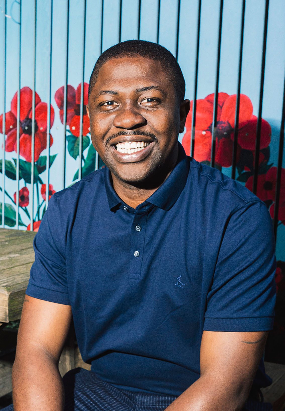 Portrait of Edafe Okporo smiling, in front of a wall of colorful red flower illustrations.