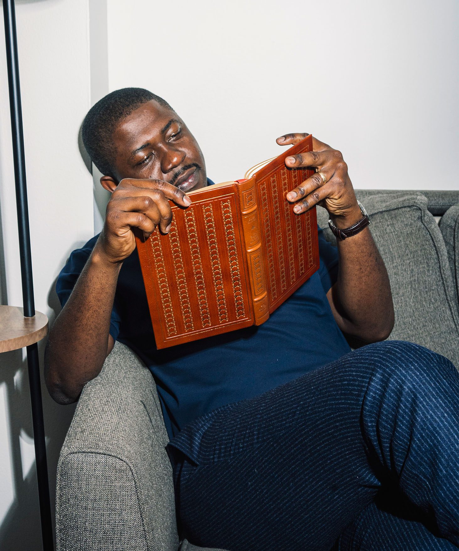 Edafe Okporo seated on a sofa, reading a book in a brown leather binding.