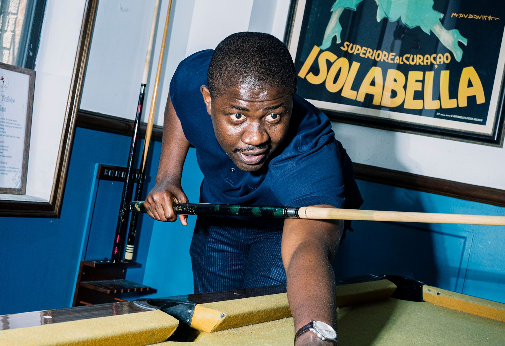Edafe Okporo playing pool. He's bending in front of a billiard table with a cue stick in his hand, looking past the camera. In the background is a poster on a wall that reads, “Isolabella.”