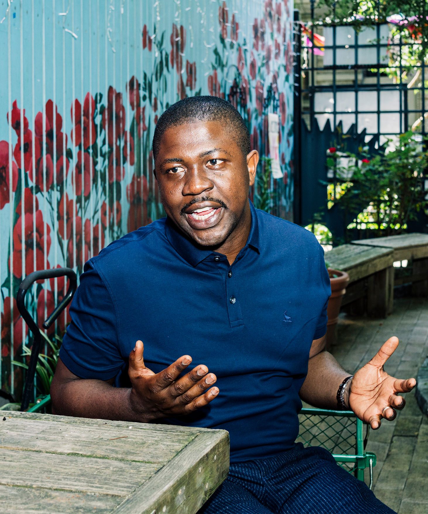 Edafe Okporo seated, in discussion with the garden and a colorful painted wall with red flowers in the background.