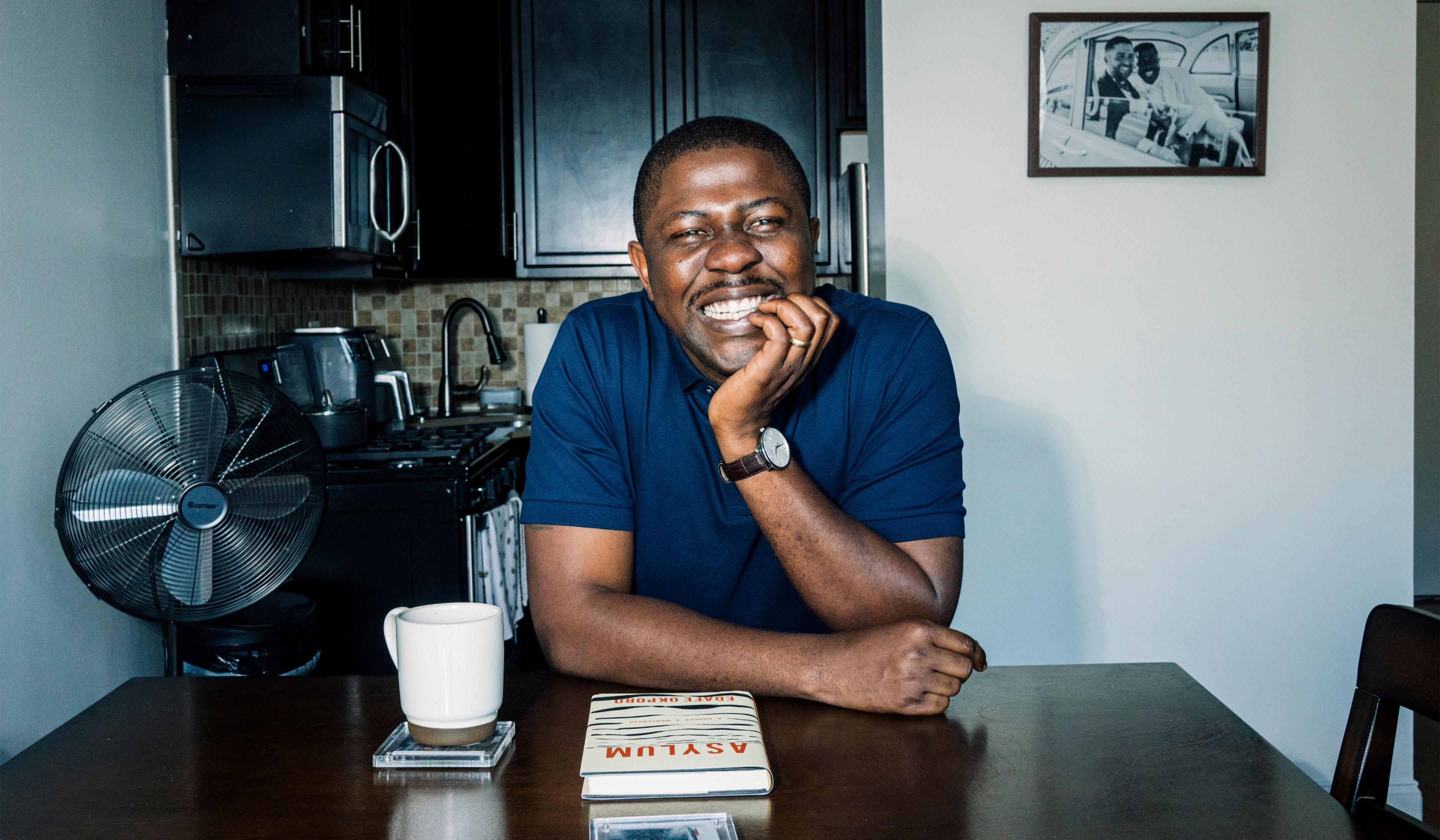 Edafe Okporo seated at a table, smiling. In front of him is a book with the title “Asylum.”