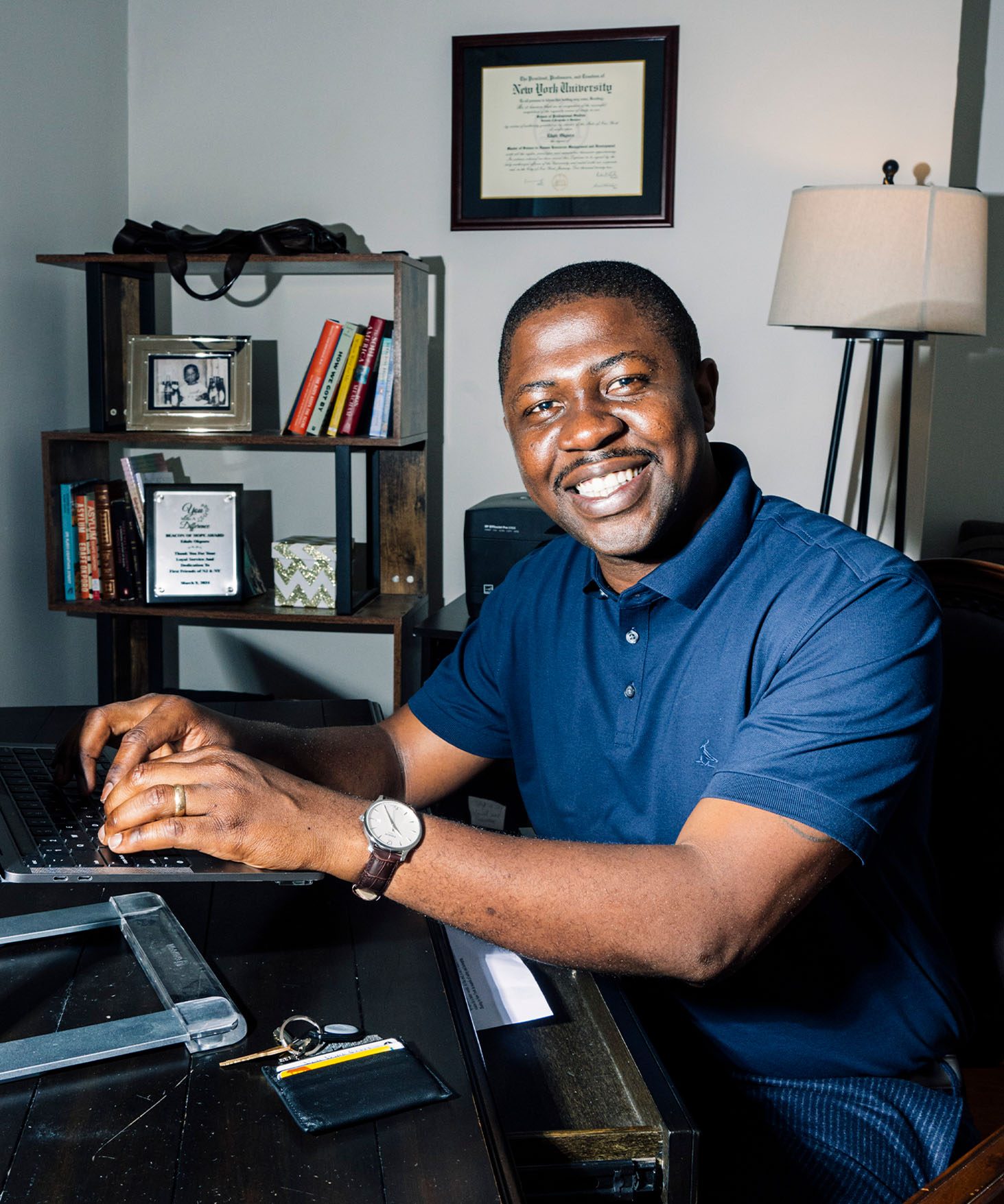 Smiling Edafe Okporo seated at a work desk. His hands are resting on a keyboard of an open laptop. A framed diploma from NYU, a lamp, and a bookshelf are visible behind him.