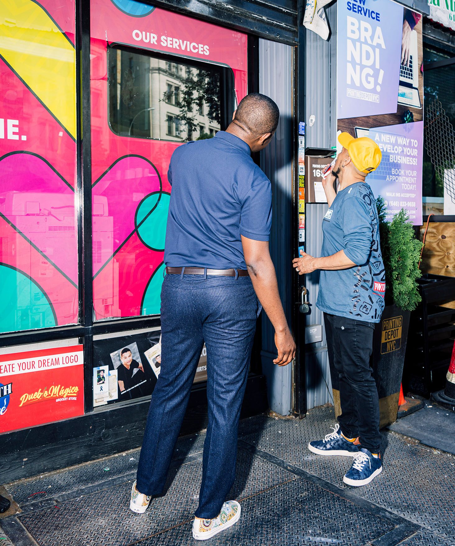 Edafe Okporo at a storefront window, with a friend from the t-shirt printing workspace. Their backs are turned away from the camera.
