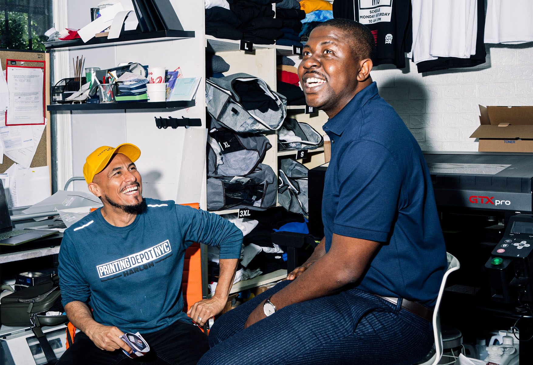 Edafe Okporo in a t-shirt printing workspace with a friend. His friend's shirt reads, “Printing Depot NYC — Harlem.”